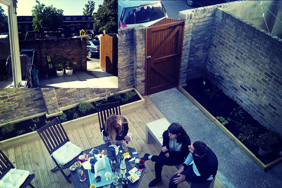 Este es un buen ejemplo de cómo un entorno puede cambiar en función del material en que está realizado. Este patio pasó de ser un entorno frío e inerte, a un espacio cálido y agradable al ser formado por maderas en su base, acompañado de unas jardineras (también en madera) y una puerta para cerrar el complejo. Todas las maderas utilizadas para la realización de este espacio son de pino macizo tratado en autoclave, con un acabado en aceite para tarimas exteriores.